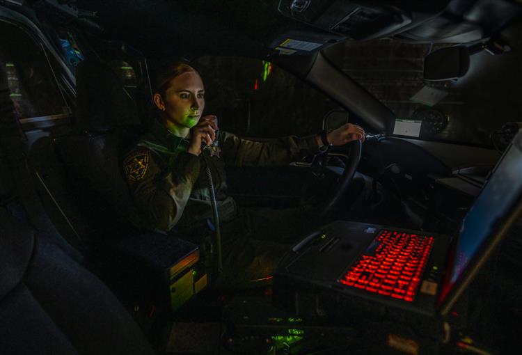 A female police officer sits in a police car at night, speaking into a radio microphone, with a laptop with a red backlit keyboard visible